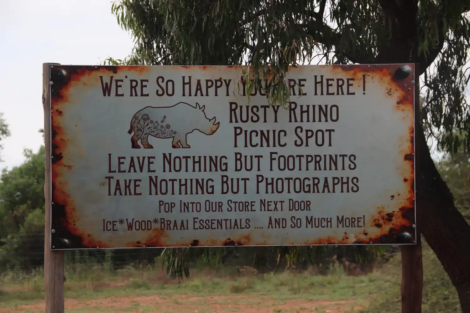 A sign that reads "We're so happy you are here! Rusty Rhino Picnic Spot. Leave nothing but footprints. Take nothing but photographs. Pop into our store next door. Ice. Wood. Braai essentials... and so much more!"